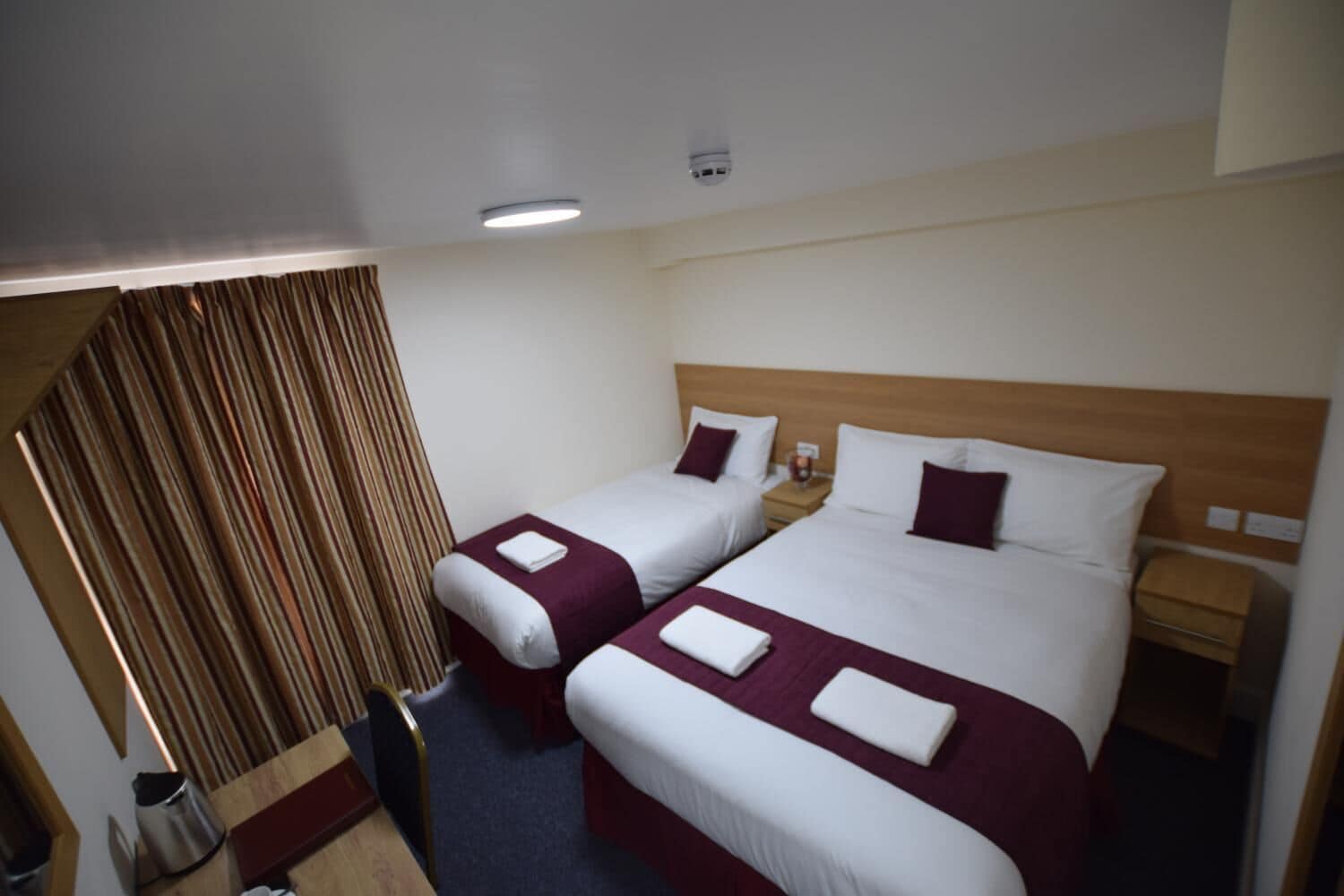 A hotel room with two beds, a wooden headboard, a nightstand, and a small table with a kettle.