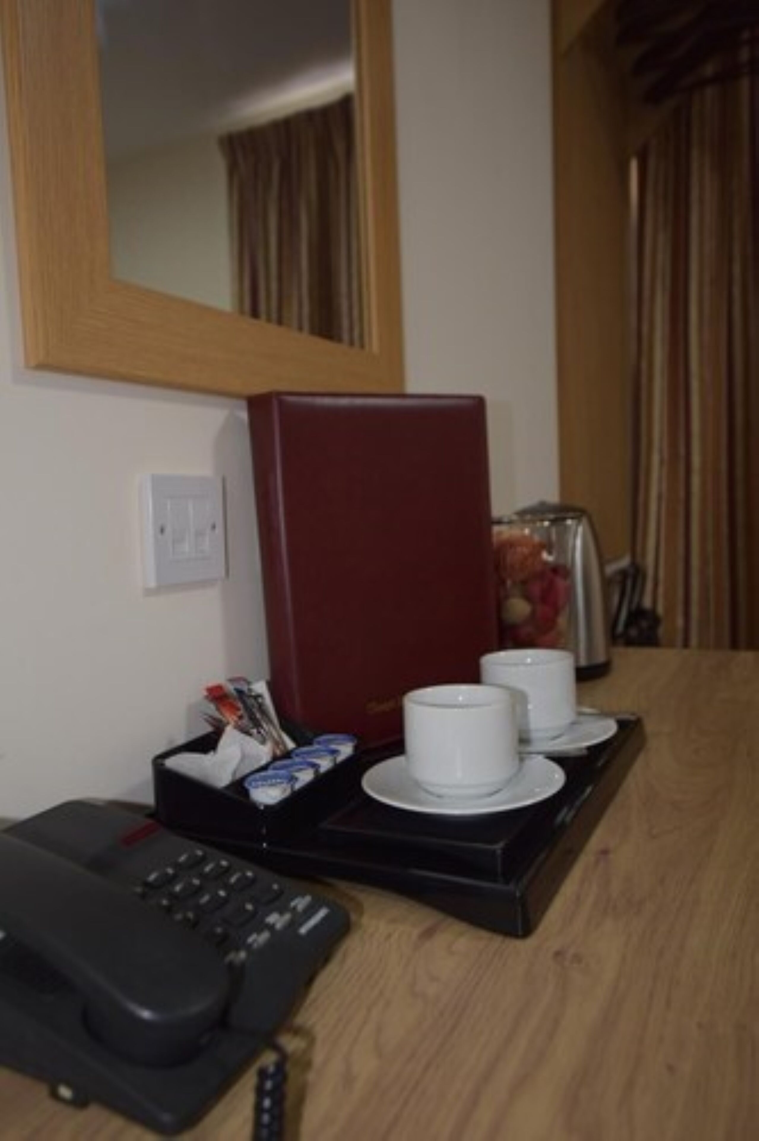 Une chambre d’hôtel avec une table en bois, un téléphone, un mini-bar rouge et un miroir au mur.