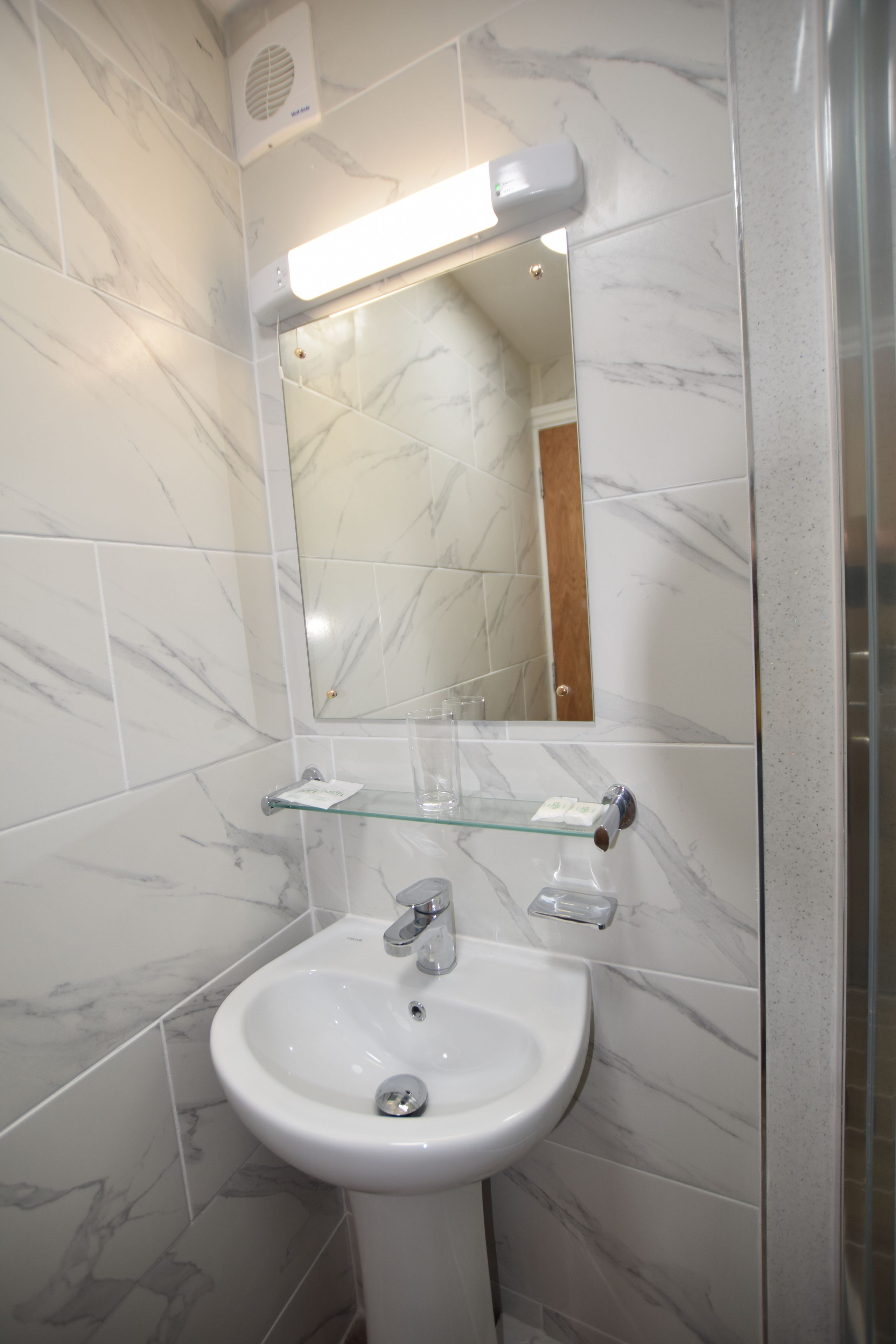 Un baño con un lavamanos de pedestal blanco, un estante de vidrio sobre el lavamanos y un espejo con luz encima.