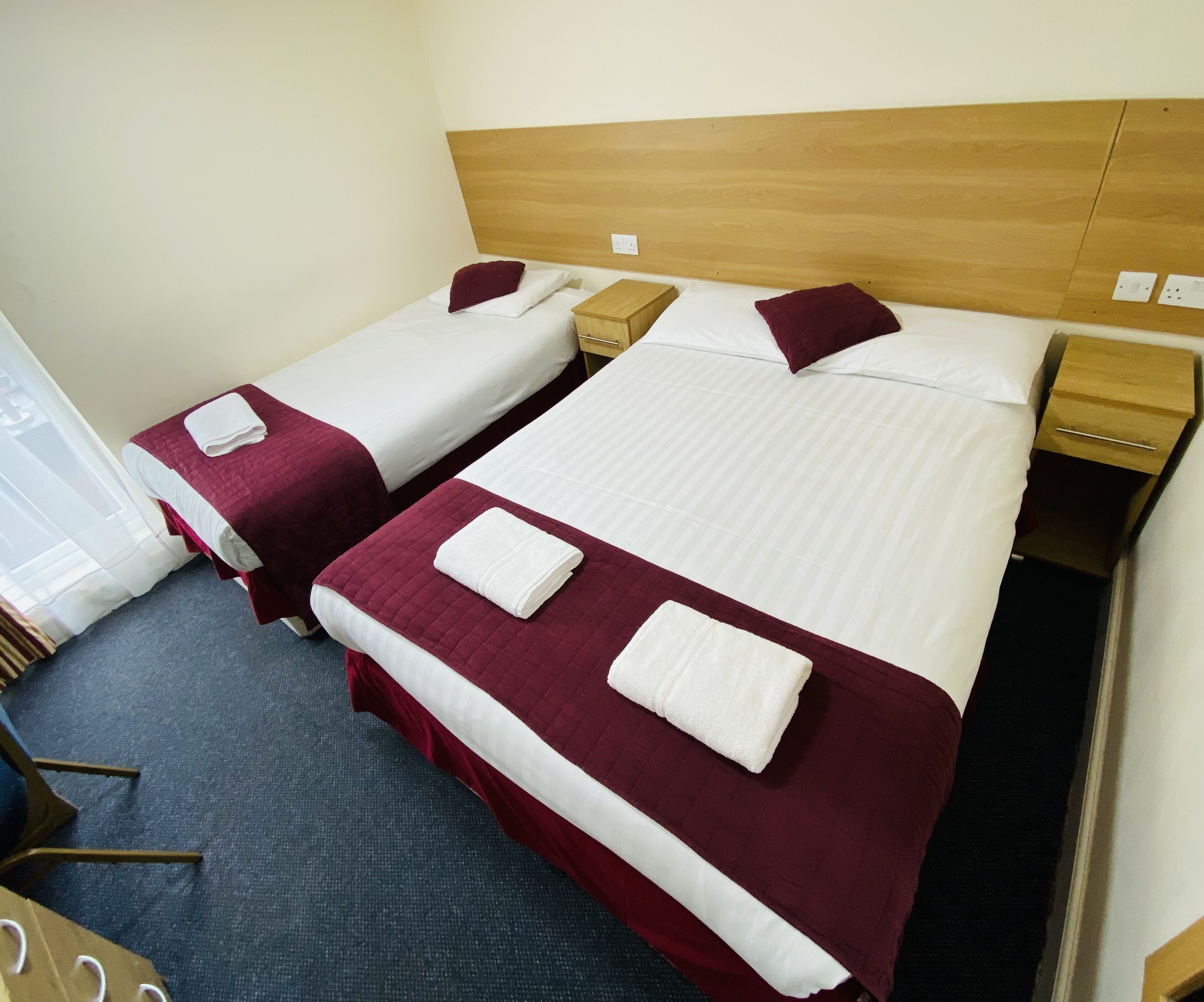 Hotellihuoneessa on kaksi erillistä sänkyä, joissa on puinen pääty, valkoiset vuodevaatteet ja tummanpunainen peitto. Huoneessa on myös puinen yöpöytä, jossa on lamppu ja pieni laatikko.