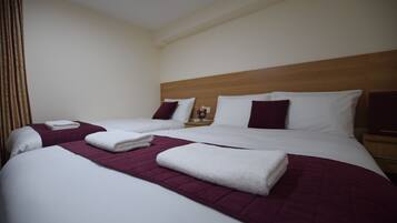 A hotel room with two beds, each with white linens and maroon bedspreads. There are two nightstands with lamps and a small table with a glass and a candle.