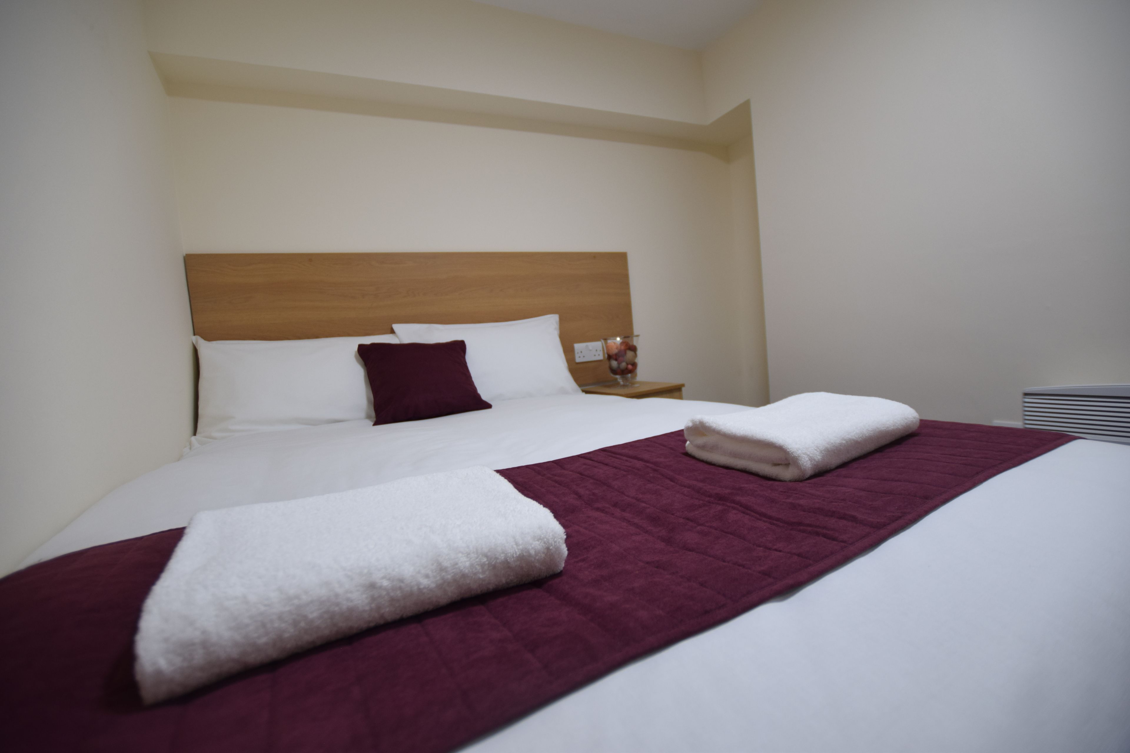A neatly made bed with a wooden headboard, white and maroon bedding, and a white towel folded on the bed.