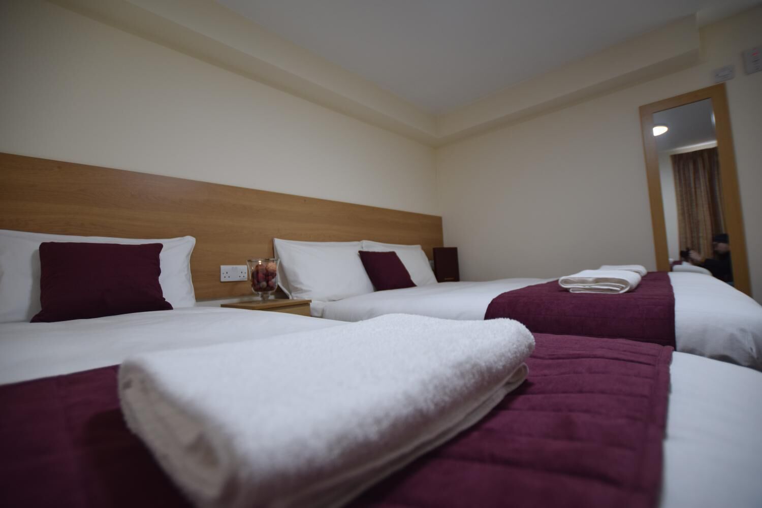 A hotel room with three beds, each with white linens and maroon pillows. There is a nightstand with a glass container and a phone.