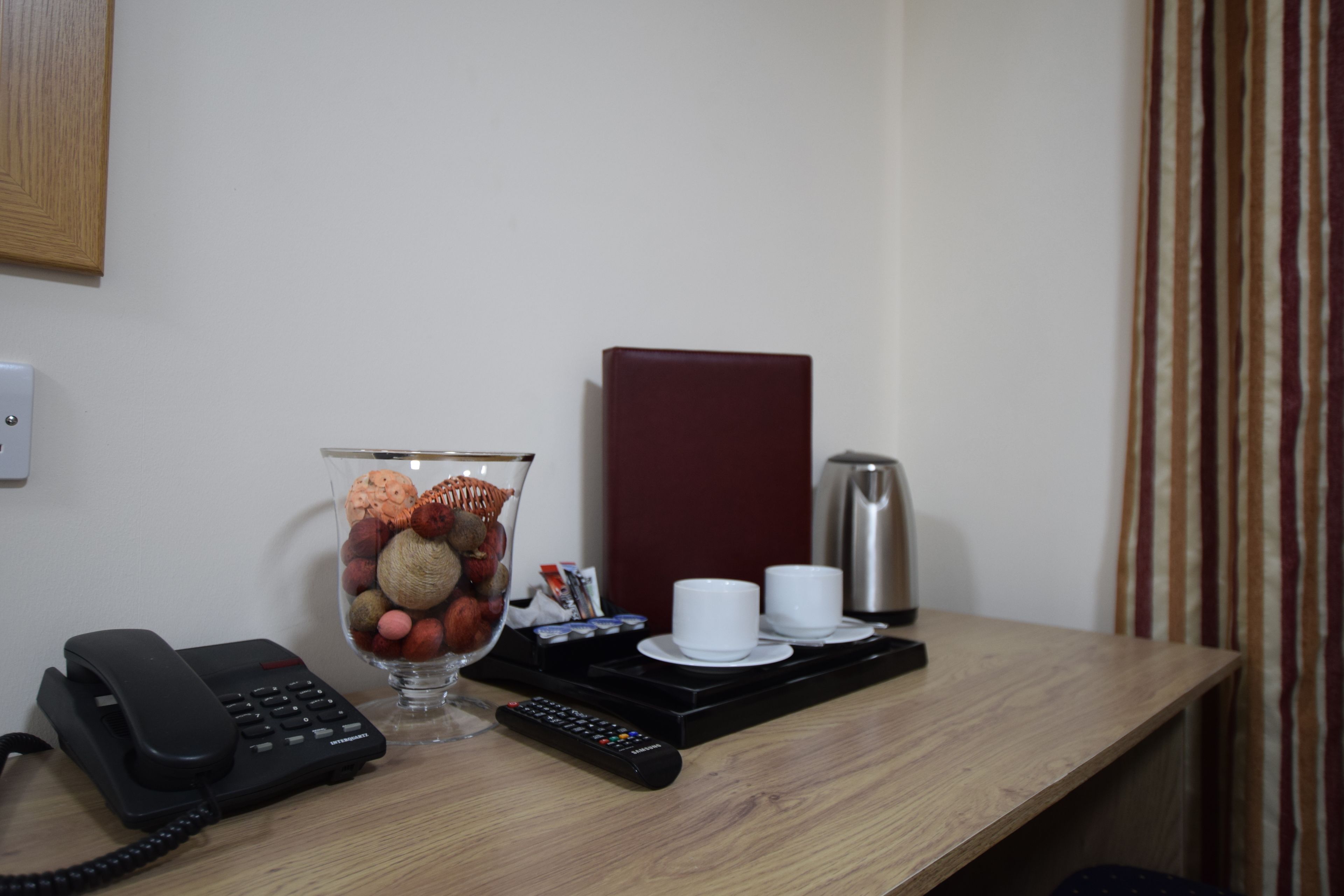 A hotel room desk with a telephone, a remote control, a glass vase with decorative items, a coffee maker, and a tray with cups and saucers.