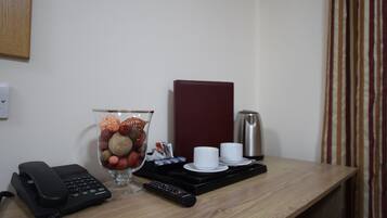 A hotel room desk with a telephone, a remote control, a glass vase with decorative items, a coffee maker, and a tray with cups and saucers.