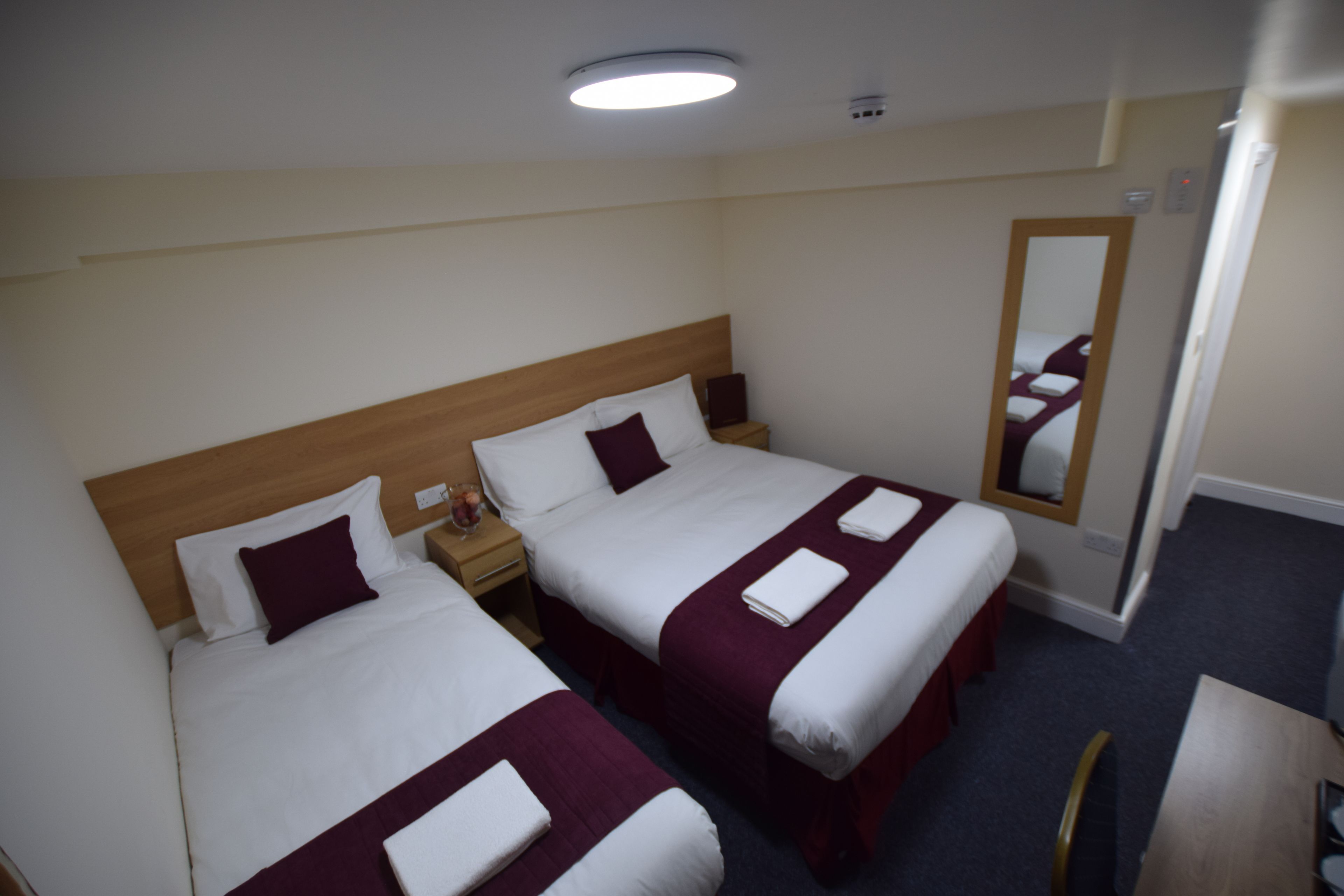 A hotel room with two beds, each with white linens and maroon accents, a wooden headboard, a nightstand with a lamp, and a mirror on the wall.