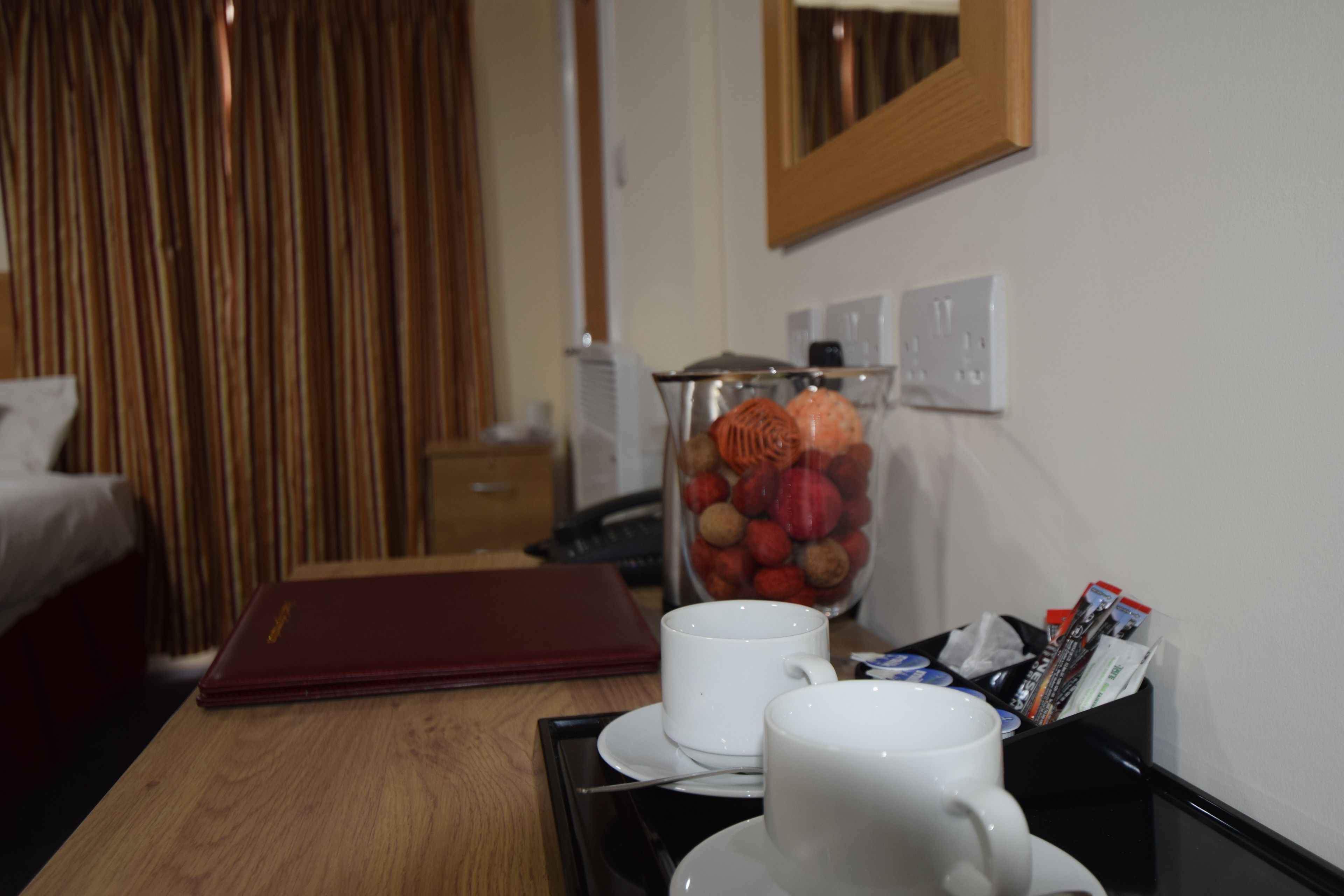 A hotel room with a wooden table, a white mug, a red notebook, a glass jar with fruits, and a black tray with pens and a phone.