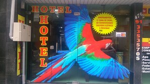 L’entrée d’un hôtel est agrémentée d’une fresque murale représentant des oiseaux aux couleurs vives, d’un panneau indiquant les équipements disponibles, comme des chambres climatisées et un réfrigérateur, et d’un numéro de téléphone bien visible.