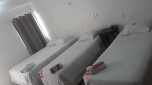 Une chambre d’hôtel avec trois lits simples, chacun recouvert d’une courtepointe blanche et muni d’une étiquette rouge. On y trouve une fenêtre avec des rideaux, un interrupteur fixé au mur et une bouche d’aération au plafond.