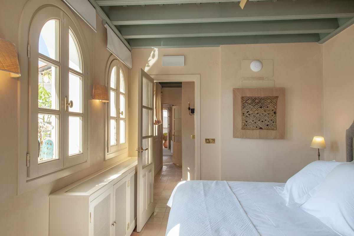 A hotel room with a large bed, a window with a view, a door leading to another room, and a wall-mounted lamp.