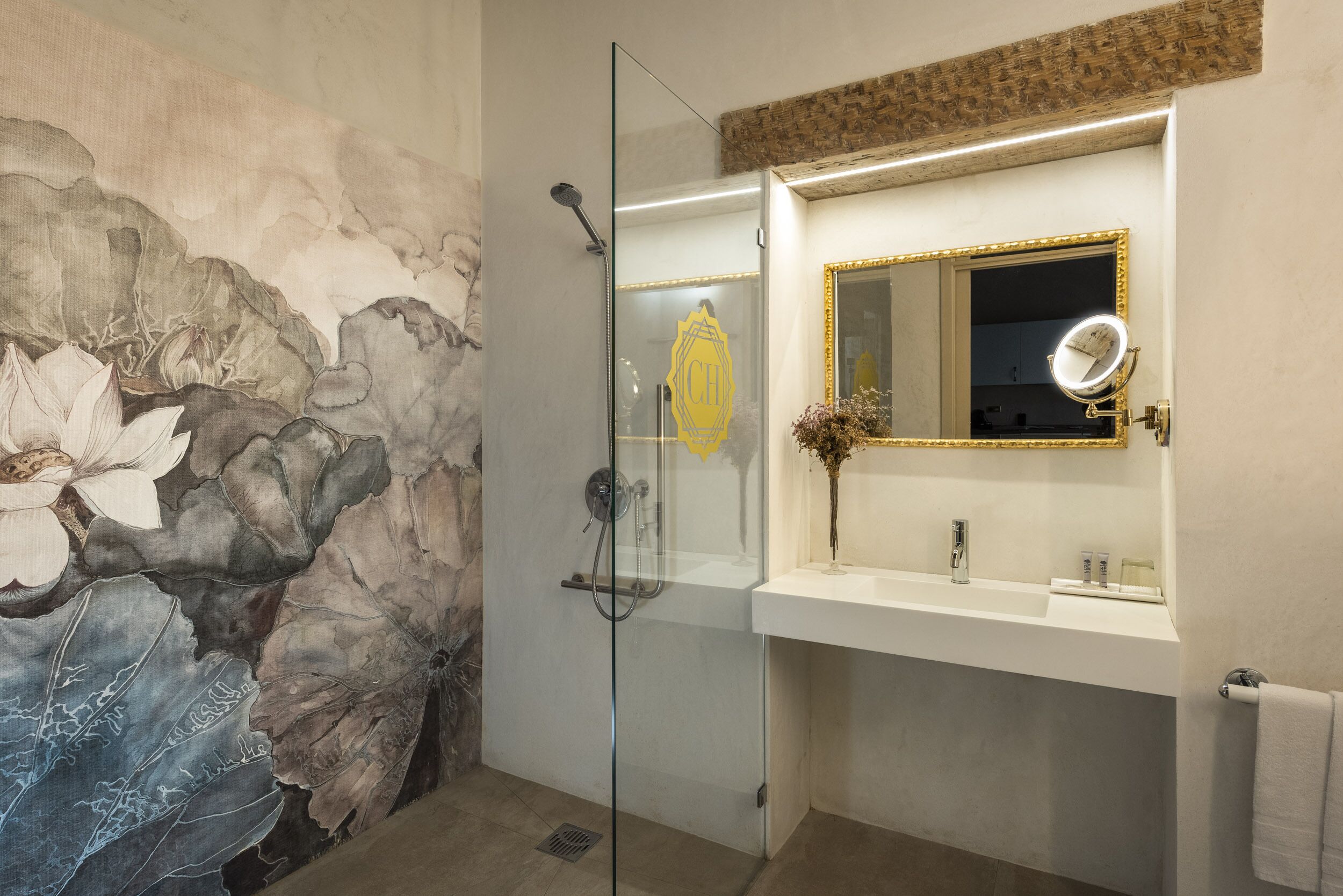 Un baño con cabina de ducha de vidrio, un lavamanos blanco con espejo de marco dorado y un mural floral en la pared.