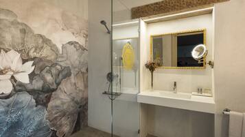 Un baño con cabina de ducha de vidrio, un lavamanos blanco con espejo de marco dorado y un mural floral en la pared.