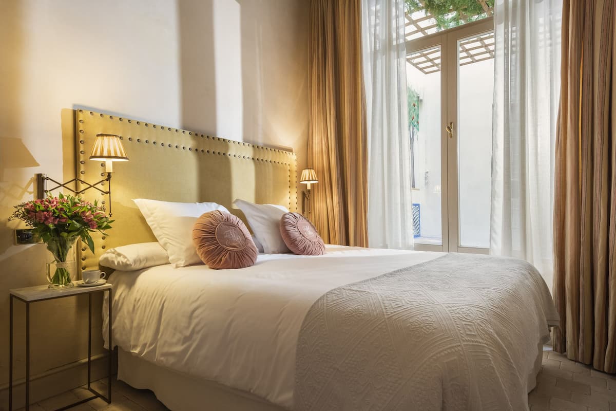 A hotel room with a large bed, two bedside lamps, a vase with flowers, and a view of the outdoors through a glass door.