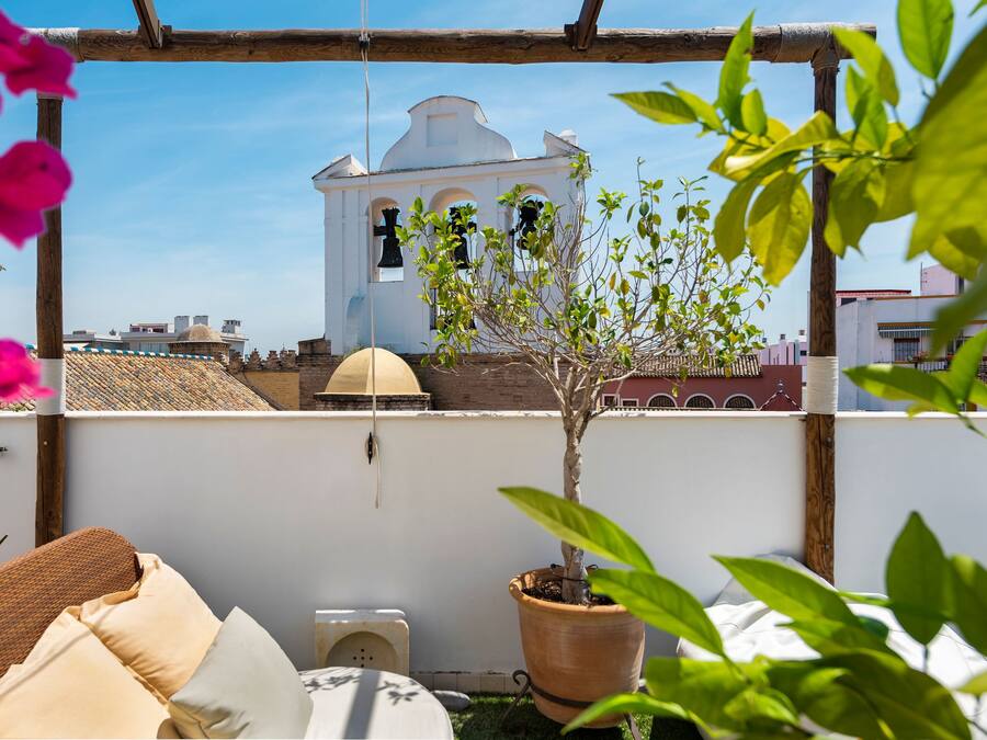 A rooftop terrace with a white bell tower, a potted tree, and a wicker sofa.