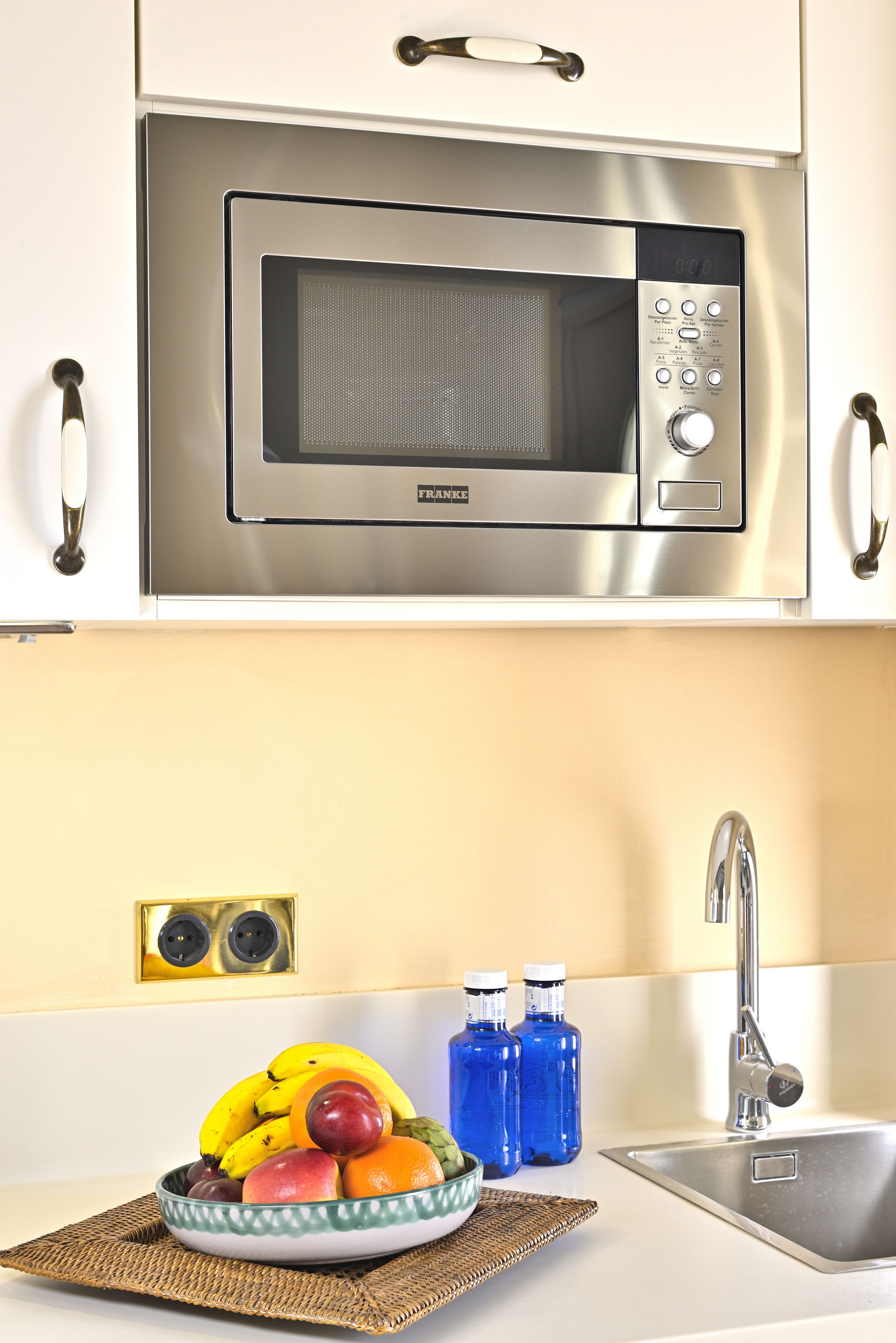 A kitchen with a stainless steel microwave, a bowl of fruit, and a sink.