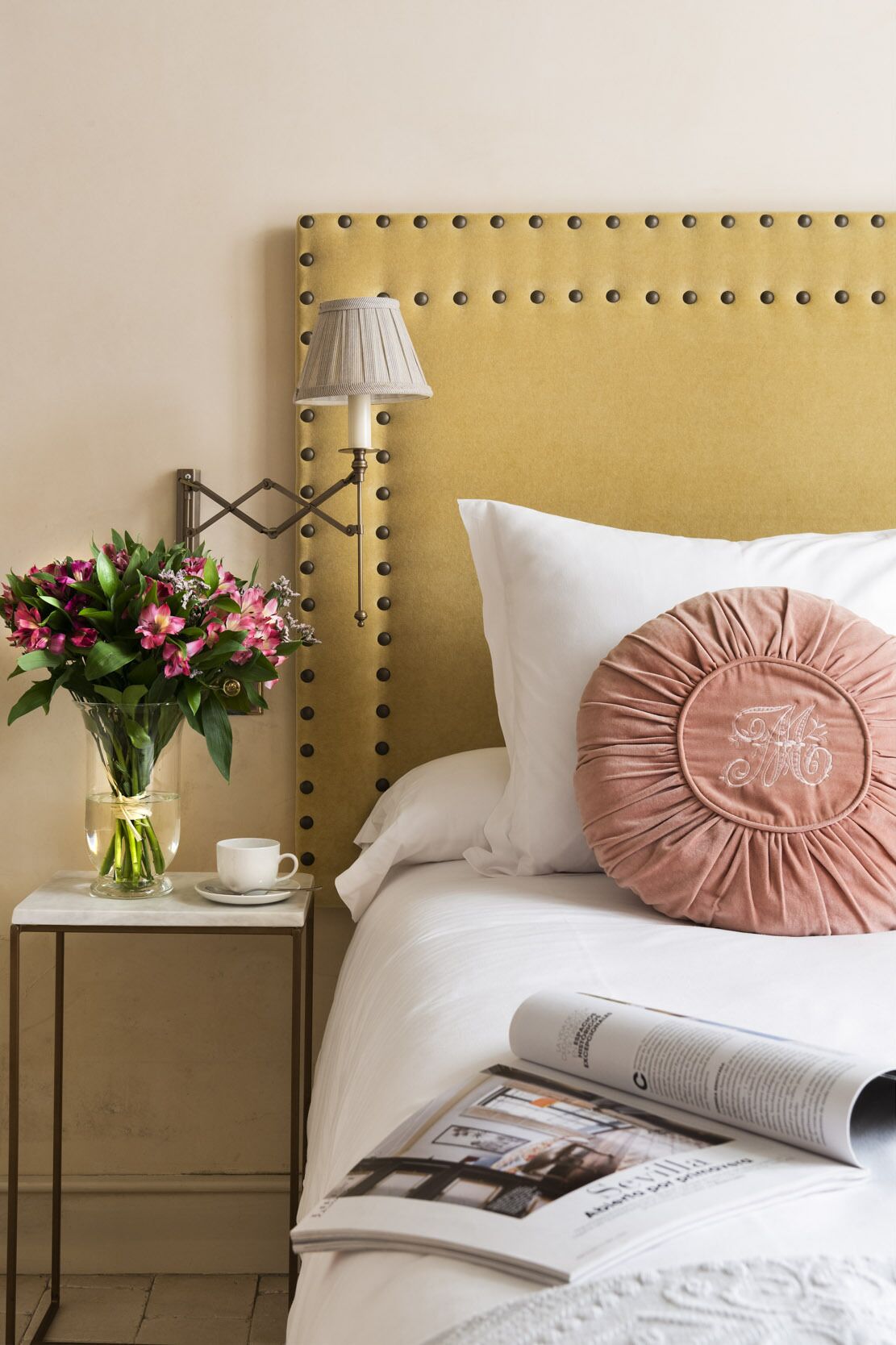 Una cama bien hecha con un cojín blanco y rosa, una revista sobre la cama, un florero con flores y una lámpara en una mesita de noche.