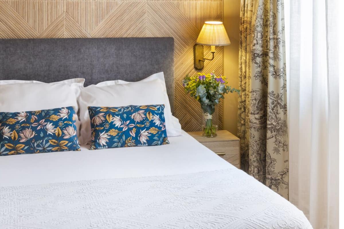 A hotel room with a patterned wall, a bed with floral pillows, a hanging light fixture, a nightstand with a lamp and flowers, and a window with curtains.