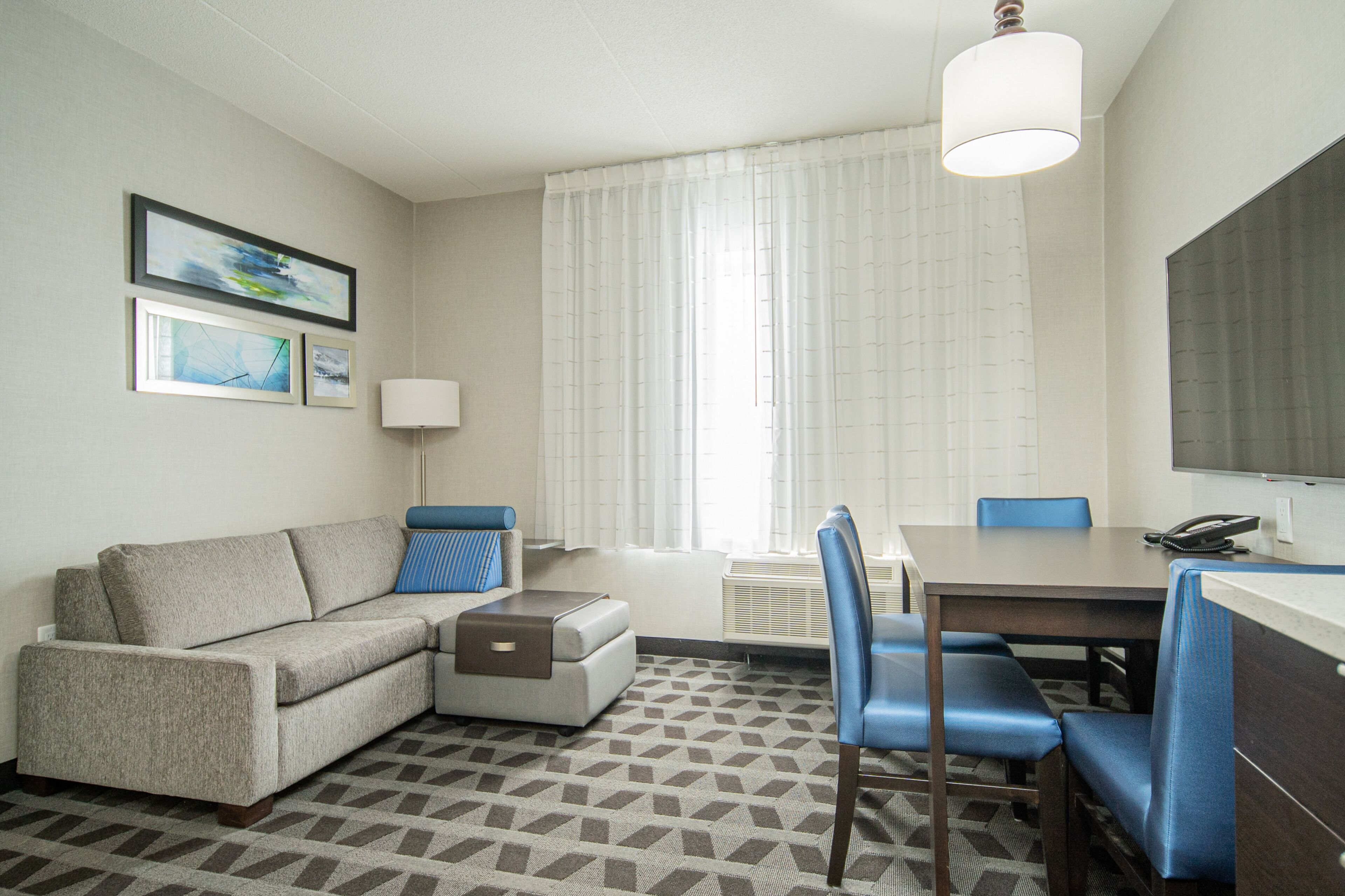 A hotel room with a sofa, ottoman, dining table, chairs, a lamp, and artwork on the wall.
