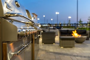 Una parrilla para barbacoa al aire libre con varios quemadores, un asa metálica y un panel de control. Se observa un brasero con llamas al fondo, y un área de estar exterior con cojines y una mesa.