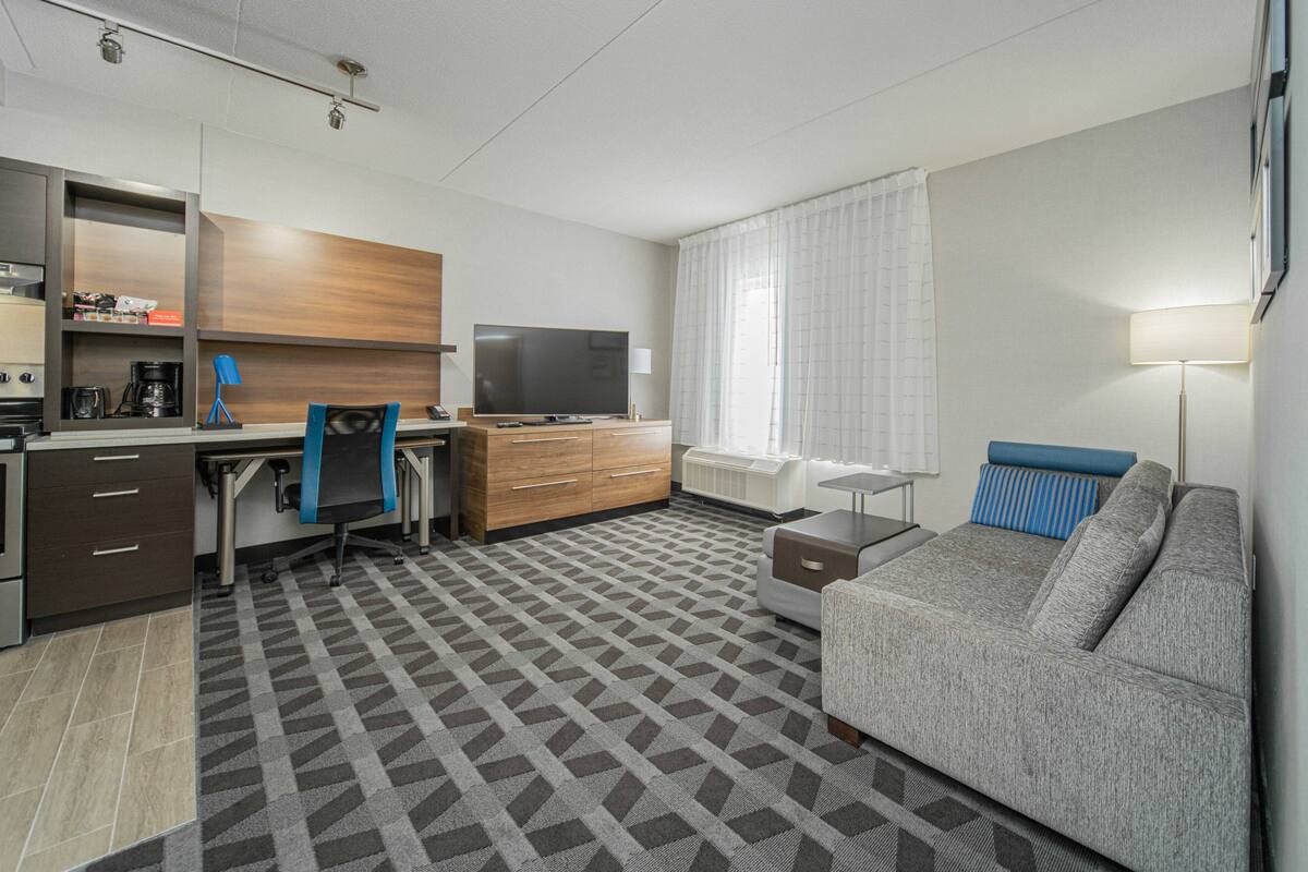 A modern hotel room with a kitchenette, a flat-screen TV, a sofa, and a patterned carpet.