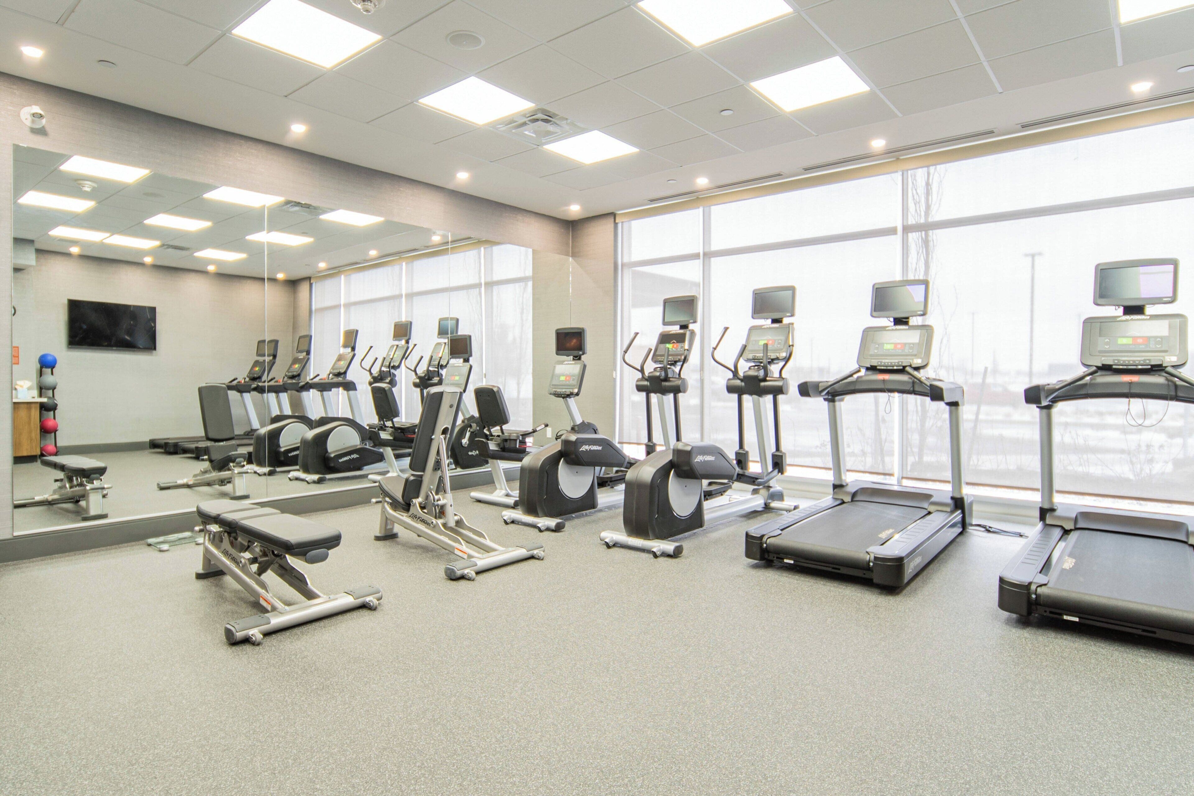 A gym with various exercise equipment, large windows, and a mirror on the wall.
