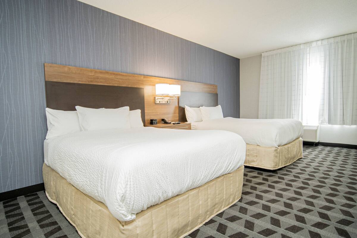 A hotel room with two beds, a wooden headboard, a nightstand with a lamp, and a patterned carpet.