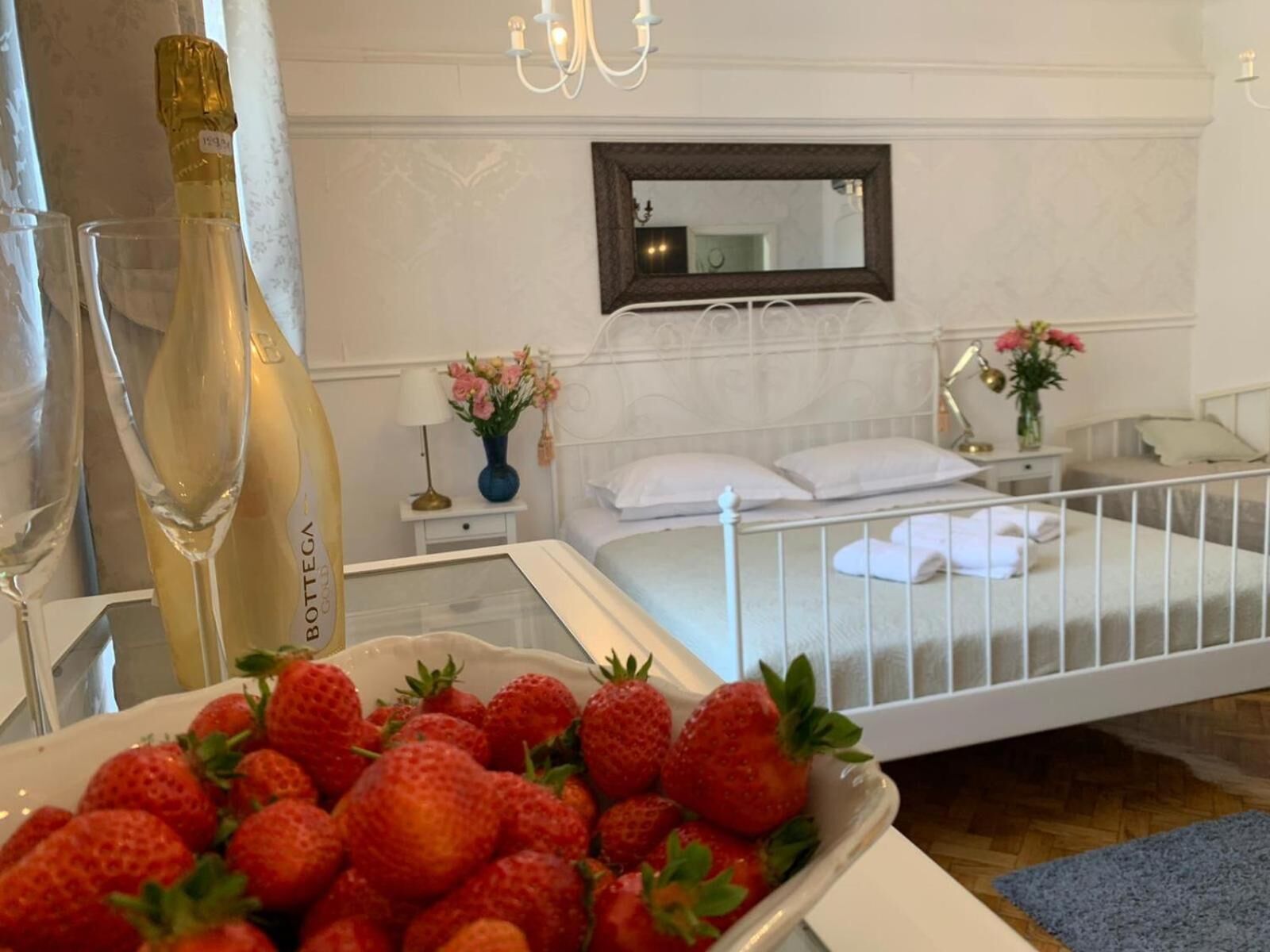 A bedroom with a bed, a nightstand, a bedside table, a mirror, and a tray with strawberries.