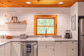 Une cuisine avec un plafond en bois, des armoires blanches, un évier, un grille-pain et une fenêtre donnant sur des arbres.