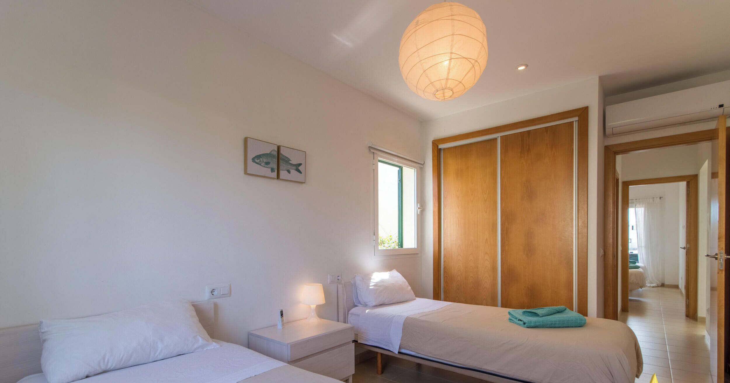 Une chambre d’hôtel avec deux lits, une armoire en bois, une table de chevet avec une lampe et une fenêtre avec des stores.