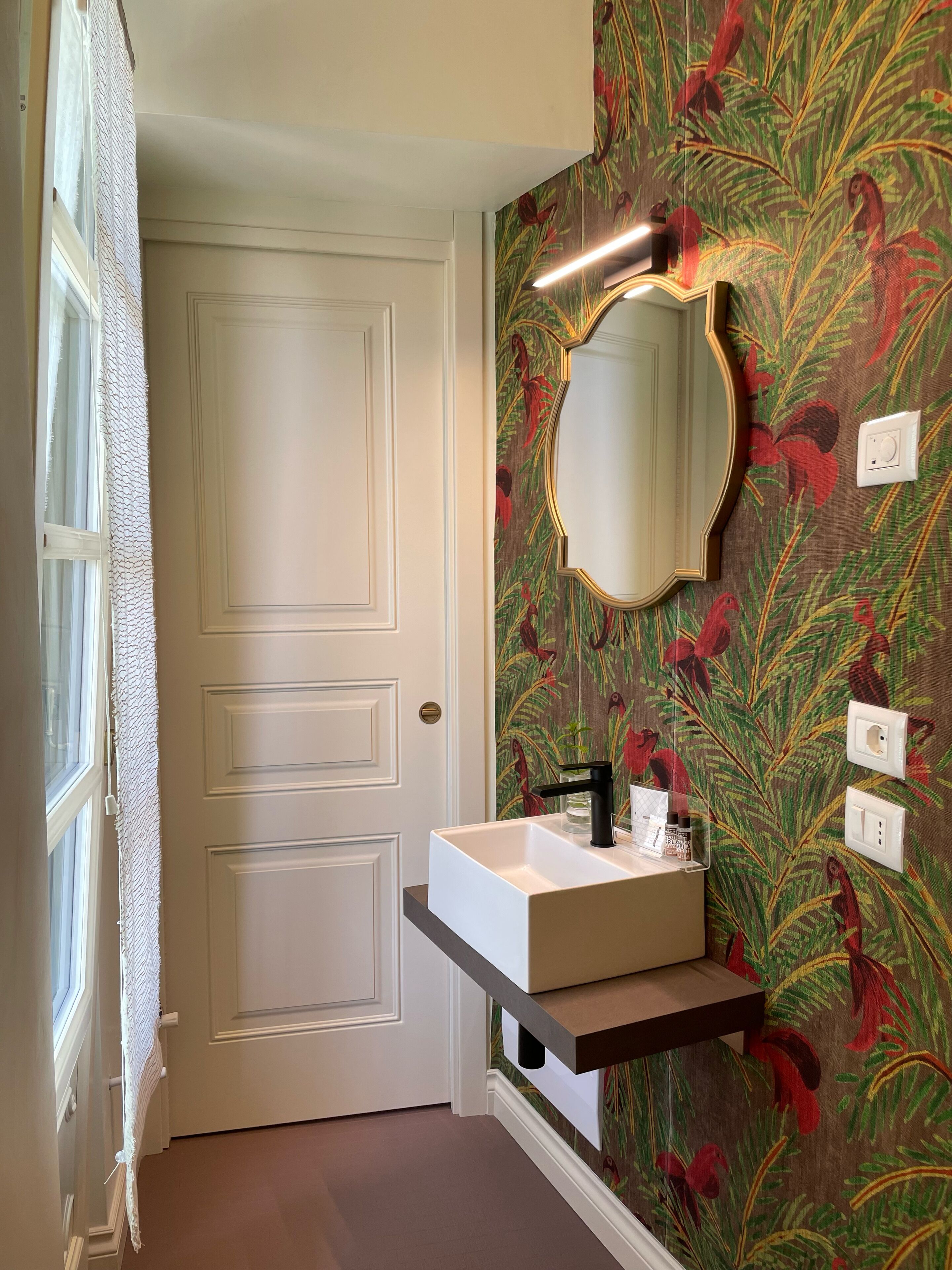 Un baño con puerta blanca, un lavamanos blanco con grifo negro, un espejo con marco dorado y una pared con estampado de hojas tropicales.