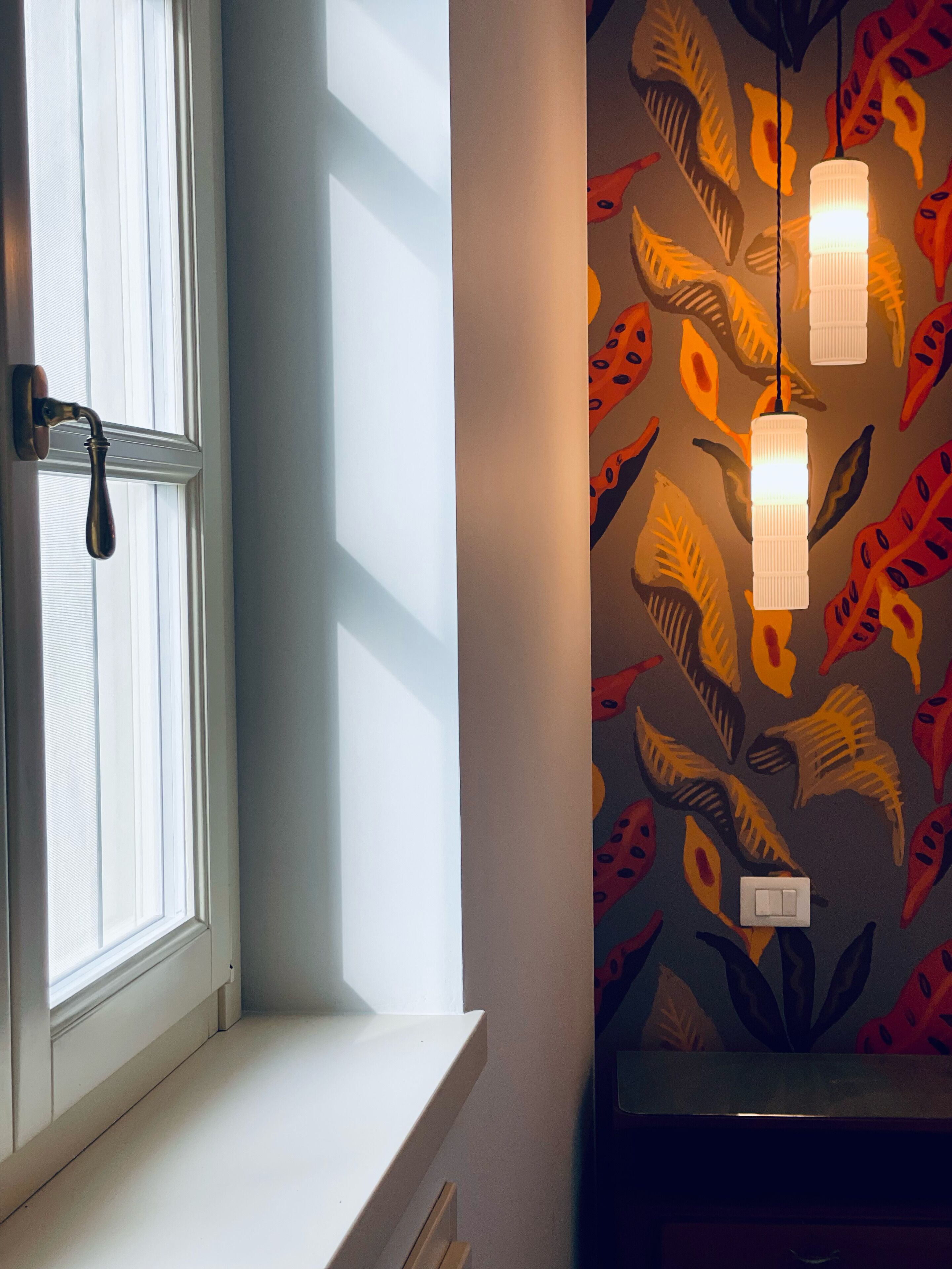 A room with a window, a wall with a colorful leaf pattern, and two hanging light fixtures.