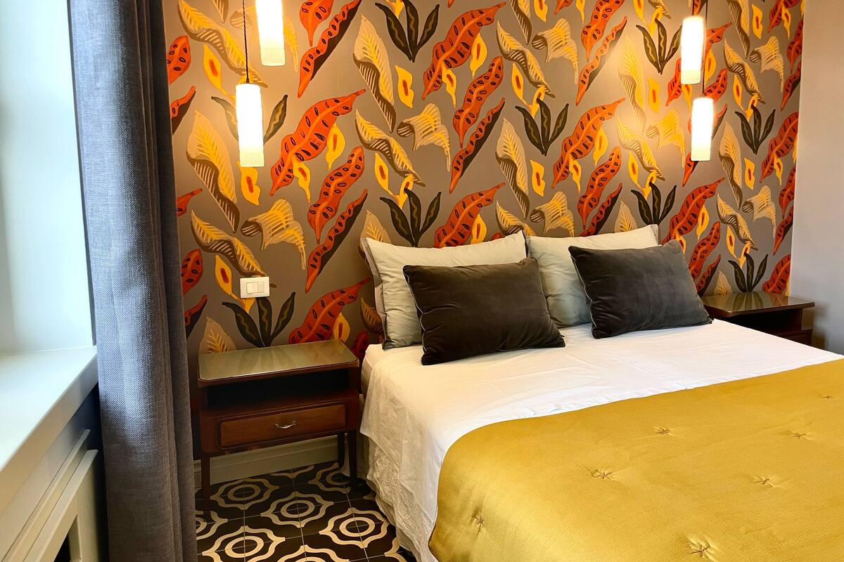 A hotel room with a patterned wallpaper, a bed with a white and gold duvet, two bedside tables, and a nightstand.