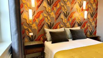 A hotel room with a patterned wallpaper, a bed with a white and gold duvet, two bedside tables, and a nightstand.