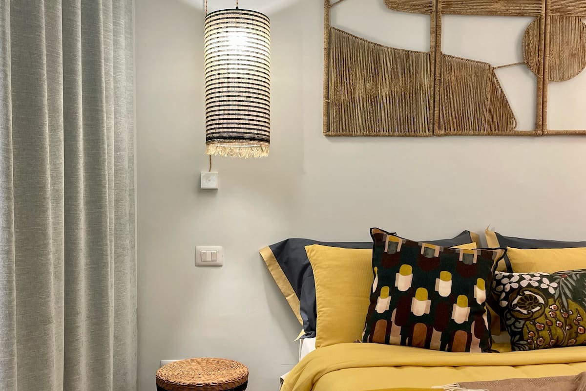 A bedroom with a bed, a hanging light fixture, a woven stool, and a wall art piece.