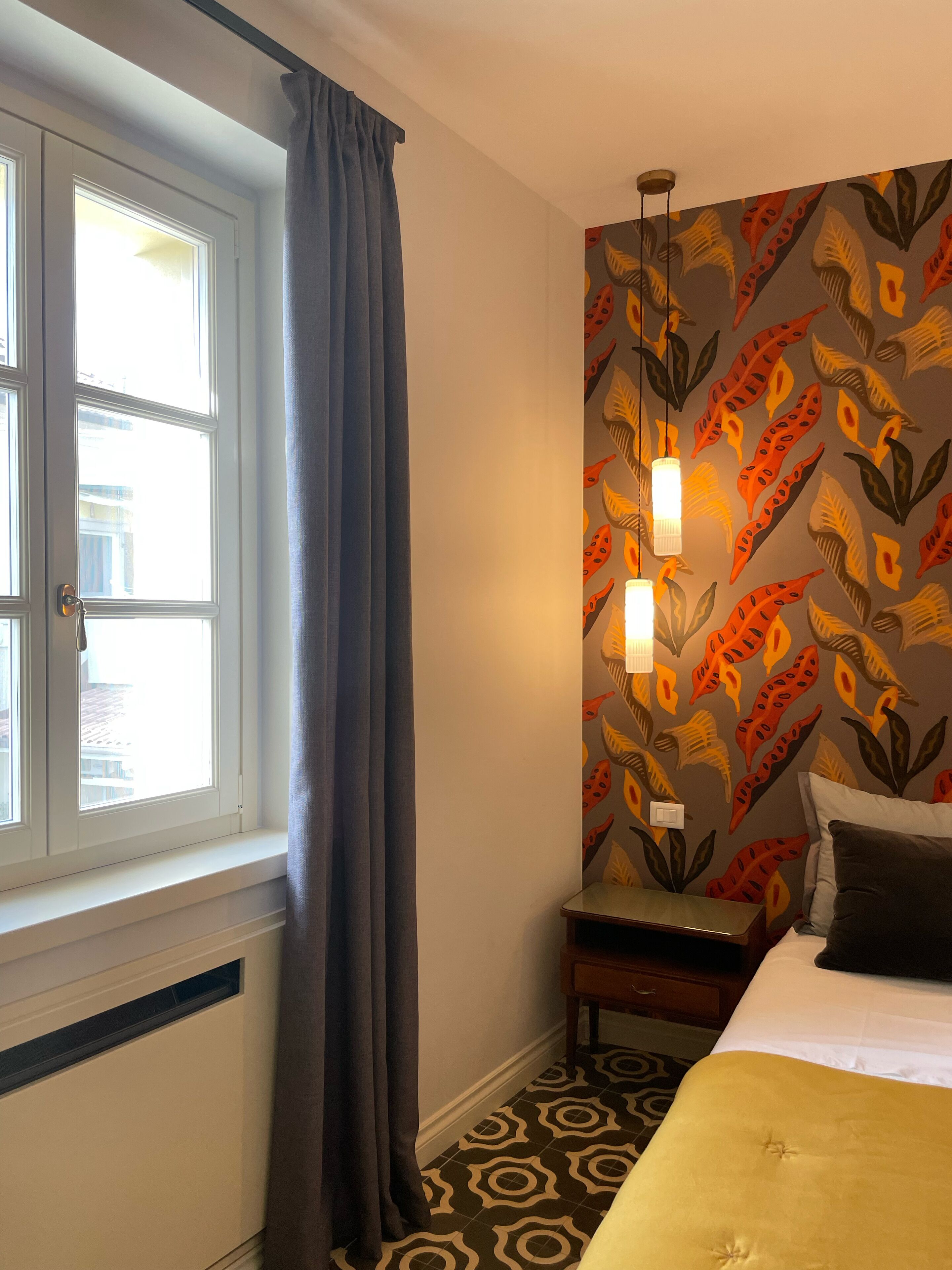 Habitación de hotel con cama, mesita de noche, lámpara, ventana con cortinas y pared con diseño.