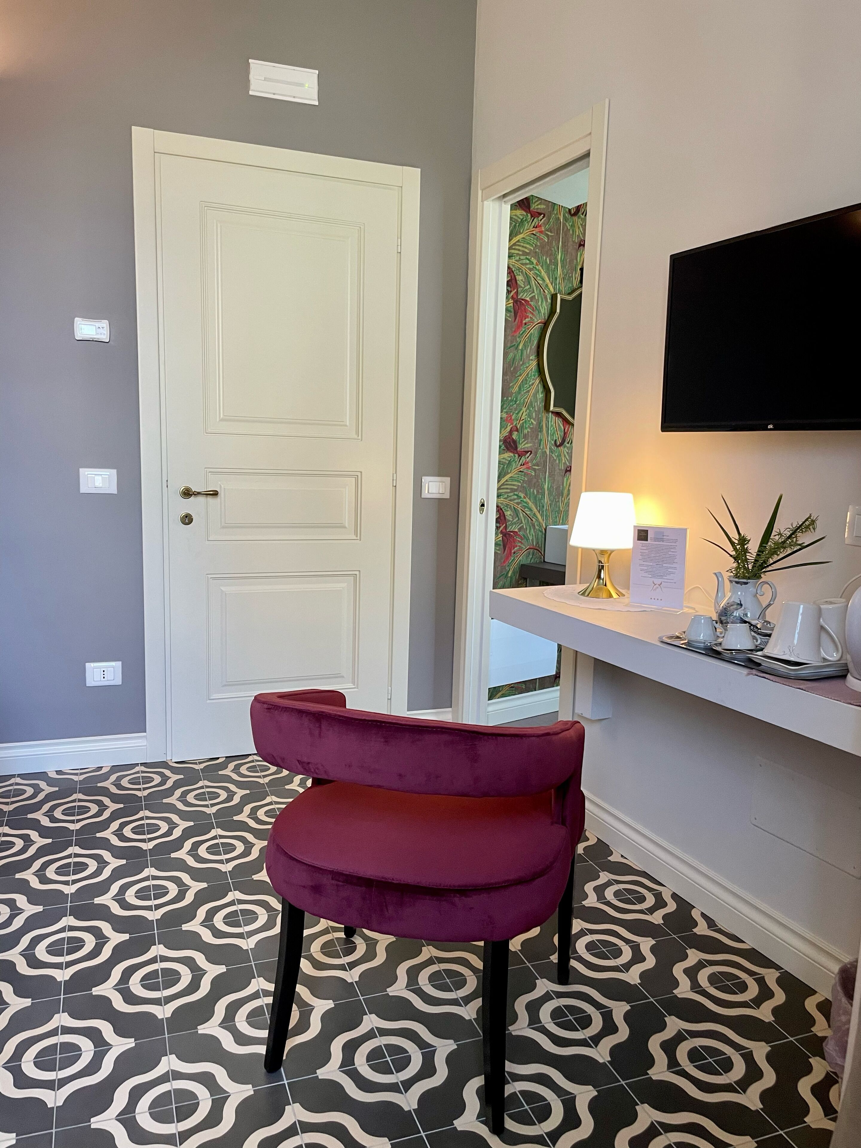 Una habitación con puerta blanca, una silla roja, un televisor de pantalla plana, una mesita con lámpara y baldosas de suelo estampadas.