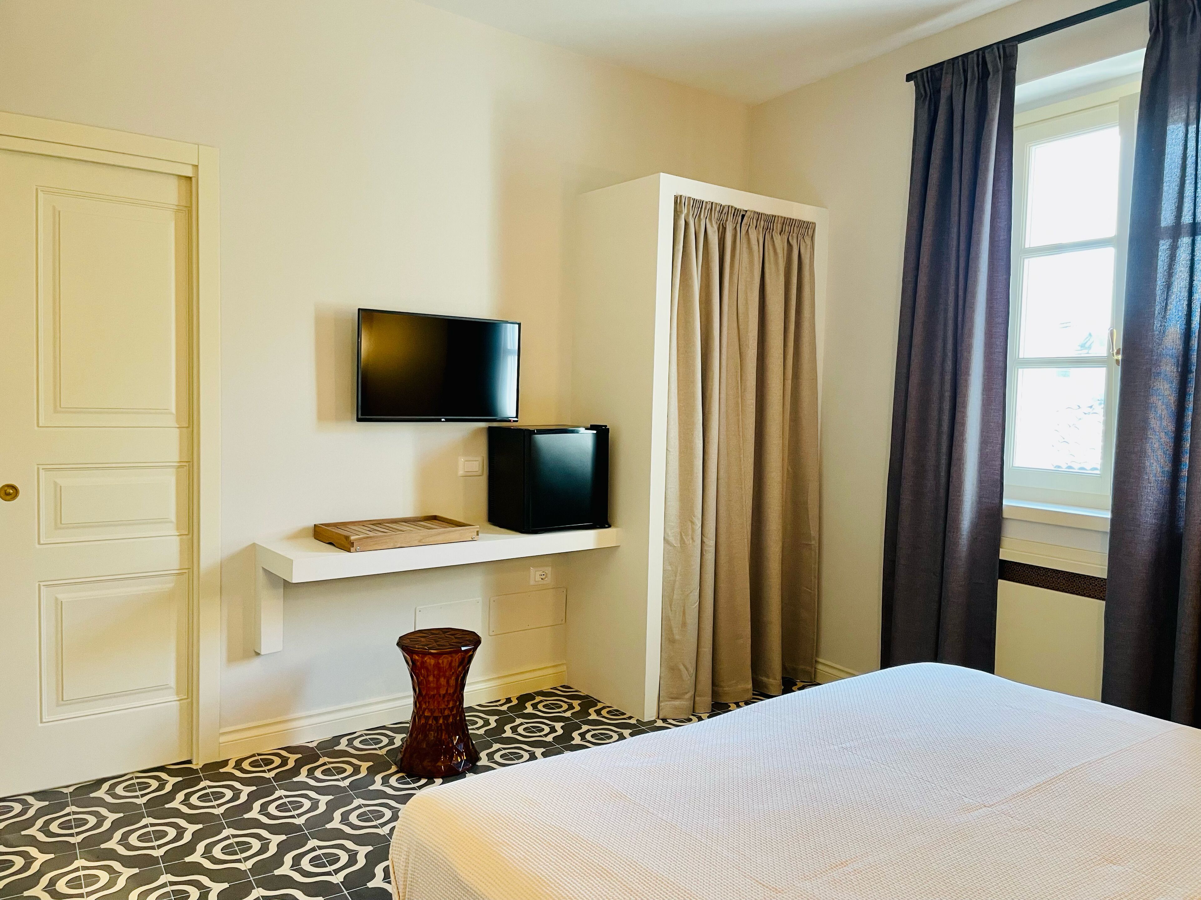 A hotel room with a bed, a TV mounted on the wall, a small shelf with a tray, a window with curtains, and patterned floor tiles.