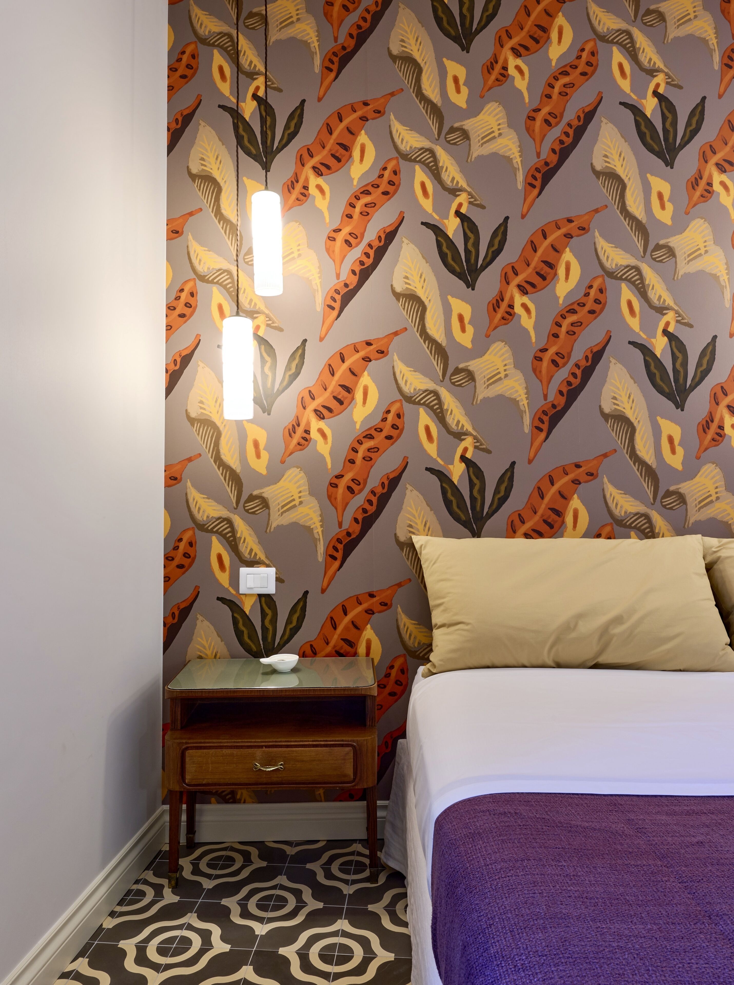A bedroom with a patterned wallpaper, a bedside table with a lamp, a bed with a purple blanket, and a patterned floor.