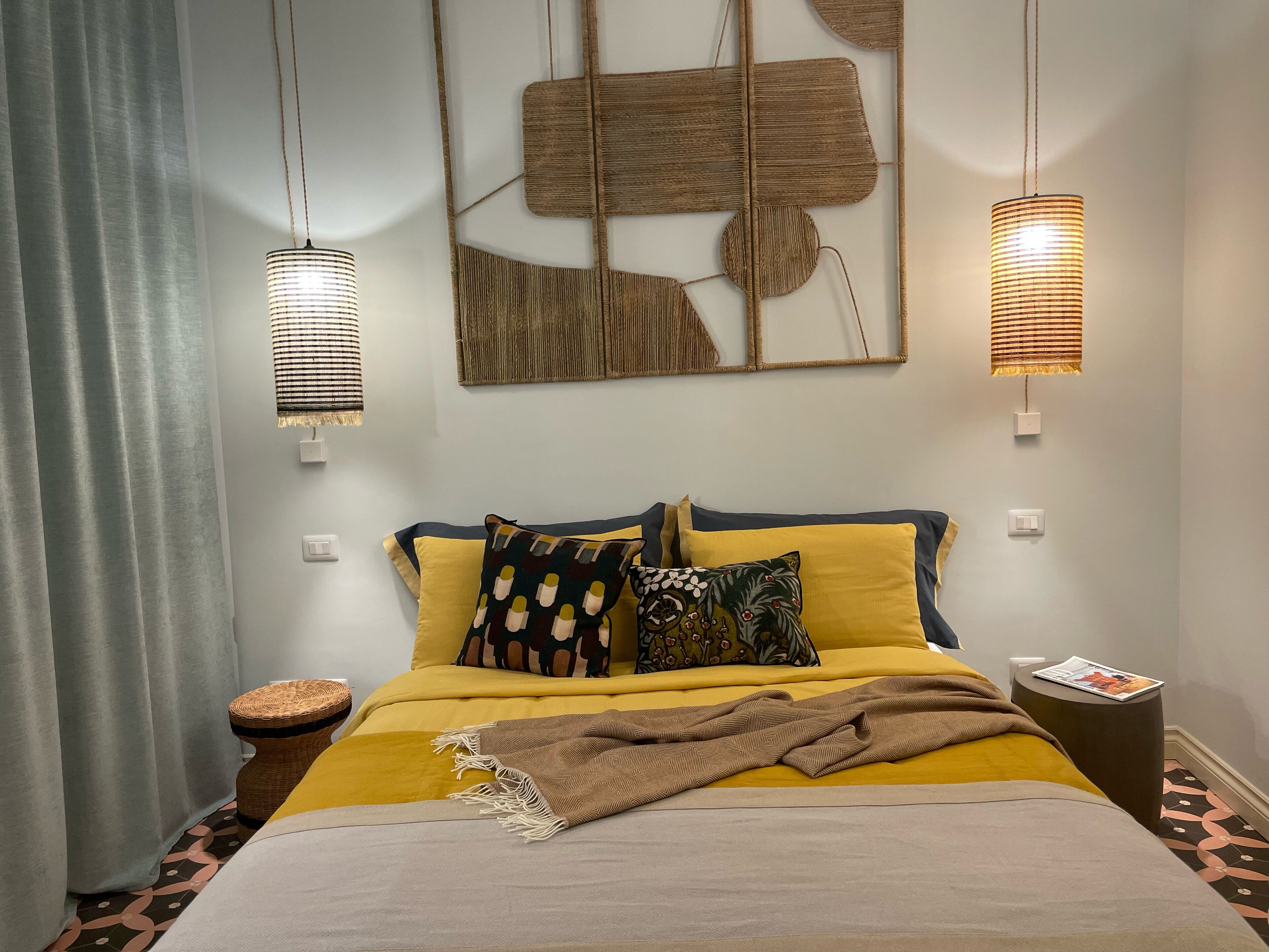 Cama bem arrumada com roupa de cama amarela, manta marrom e almofadas estampadas. Duas luminárias fixadas na parede com cúpulas listradas. Uma cesta de palha está no chão ao lado da cama.