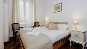 A neatly made bed with a headboard, a bedside table with a lamp, a chair, and a wall-mounted picture. - Pellegrino - WR Apartments (Rome)