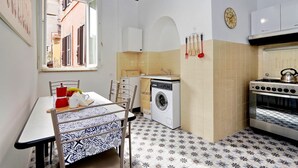 A compact kitchen with a washing machine, oven, and dining area with chairs and a table. - Pellegrino - WR Apartments (Rome)