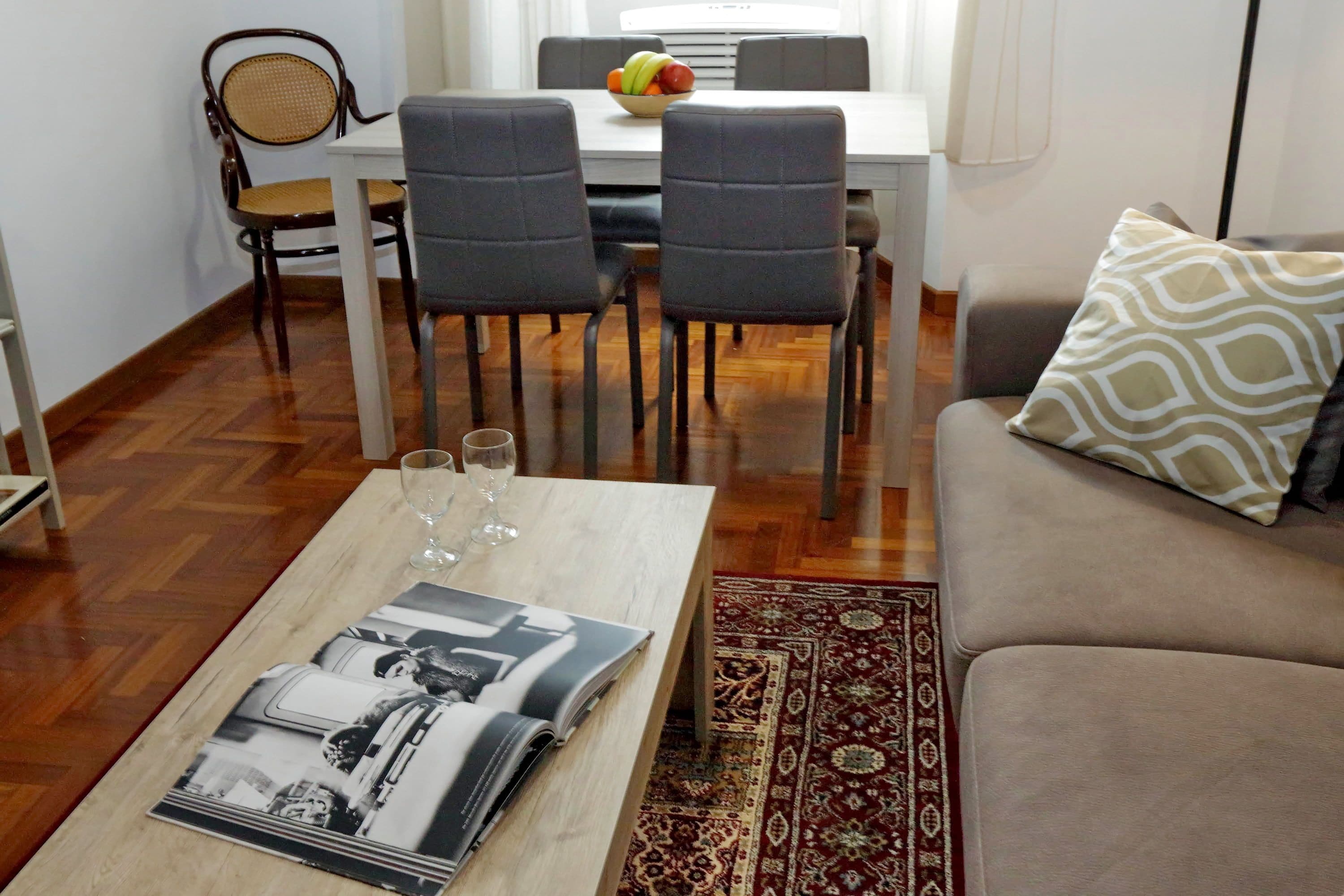 A dining area with a white table, two grey chairs, and a fruit bowl. There is a wooden coffee table with magazines and glasses. A patterned rug is on the floor.