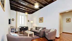 A living room with a sofa, armchair, and a coffee table. There is a dining area with a table and chairs. The room has wooden flooring and a ceiling with exposed beams. - Pellegrino - WR Apartments (Rome)