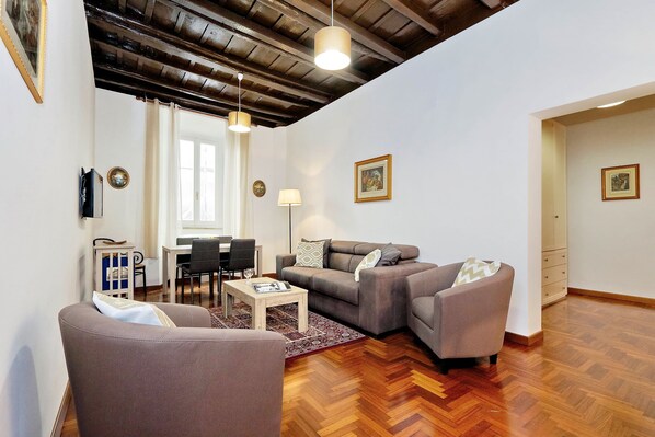 A living room with a sofa, armchair, and a coffee table. There is a dining area with a table and chairs. The room has wooden flooring and a ceiling with exposed beams.
