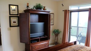 Un dormitorio con una cama grande, un mueble de madera con centro de entretenimiento y un televisor de pantalla plana, una pequeña planta en maceta y un ventanal corredero que da a un balcón.