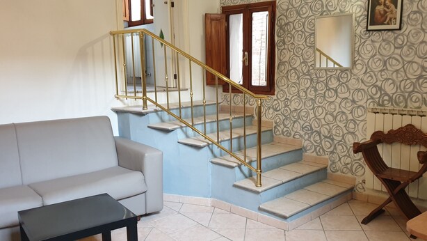 A staircase with a golden handrail, a grey sofa, a small table, and a wooden chair in a room with patterned wallpaper and a radiator. - Sapienza Time (Siena)