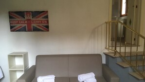 A room with a beige sofa, two rolled-up towels, a white shelf, and a staircase with a golden railing. - Sapienza Time (Siena)