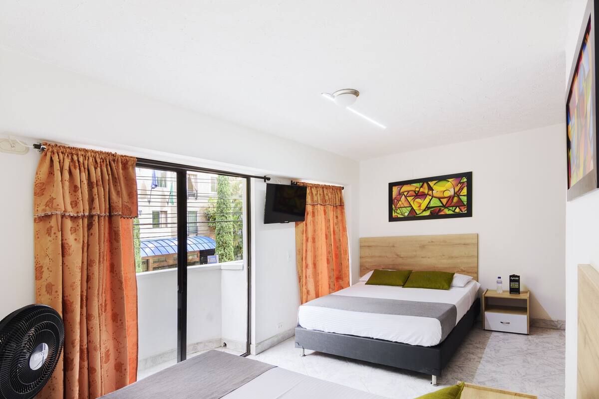 A bedroom with a bed, a nightstand, a TV, a fan, and a window with curtains.