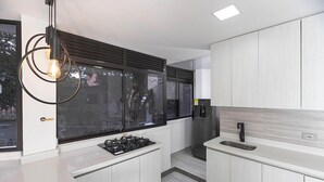 A modern kitchen with white cabinetry, a built-in gas stove, a stainless steel sink, and a pendant light with exposed bulbs. - Hotel Balcones del Estadio (Medellín)
