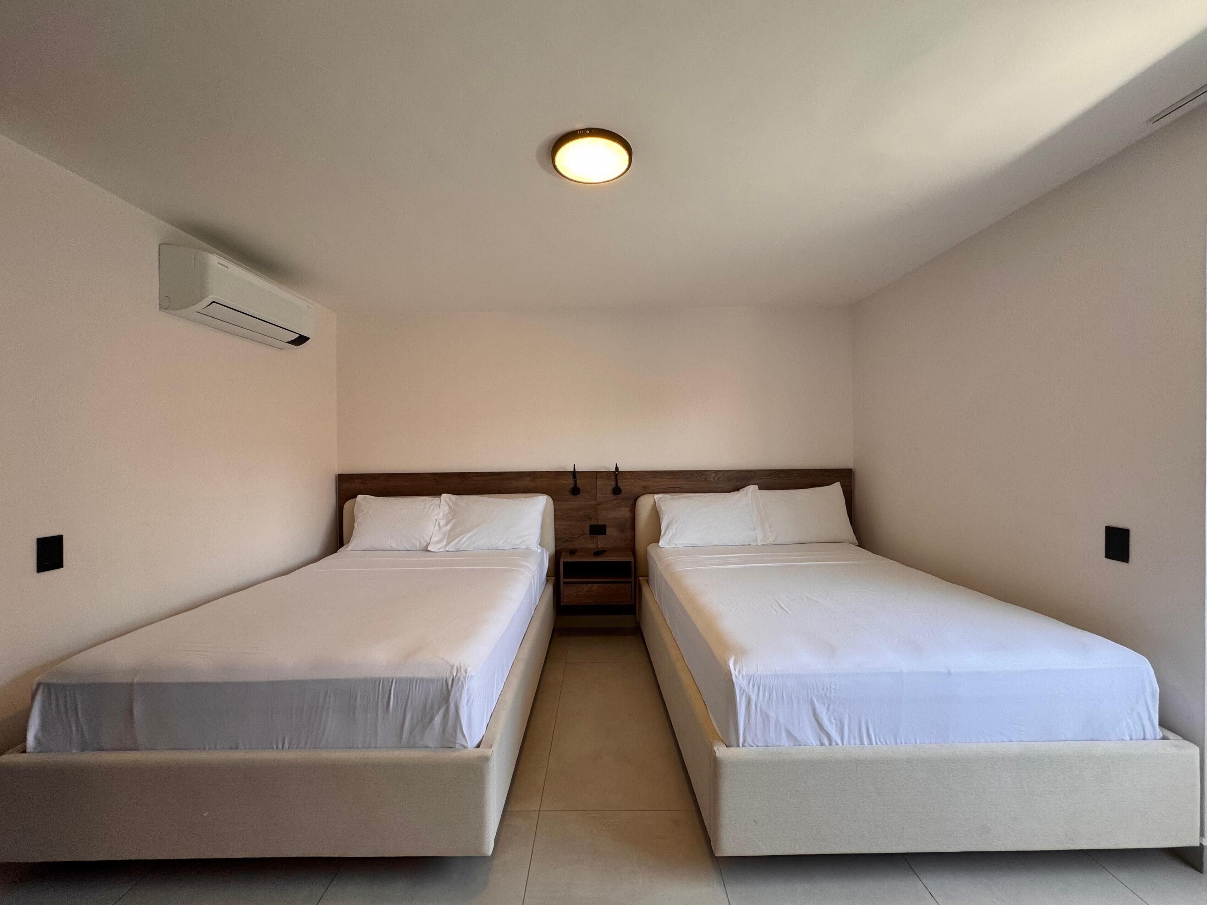 A room with two single beds, a ceiling light, and an air conditioning unit on the wall.